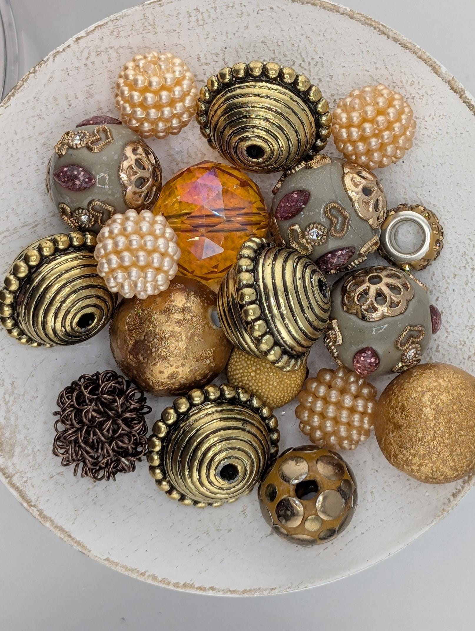 a white bowl filled with various decorative beads and ornaments, including gold, silver, and pearl-like elements.