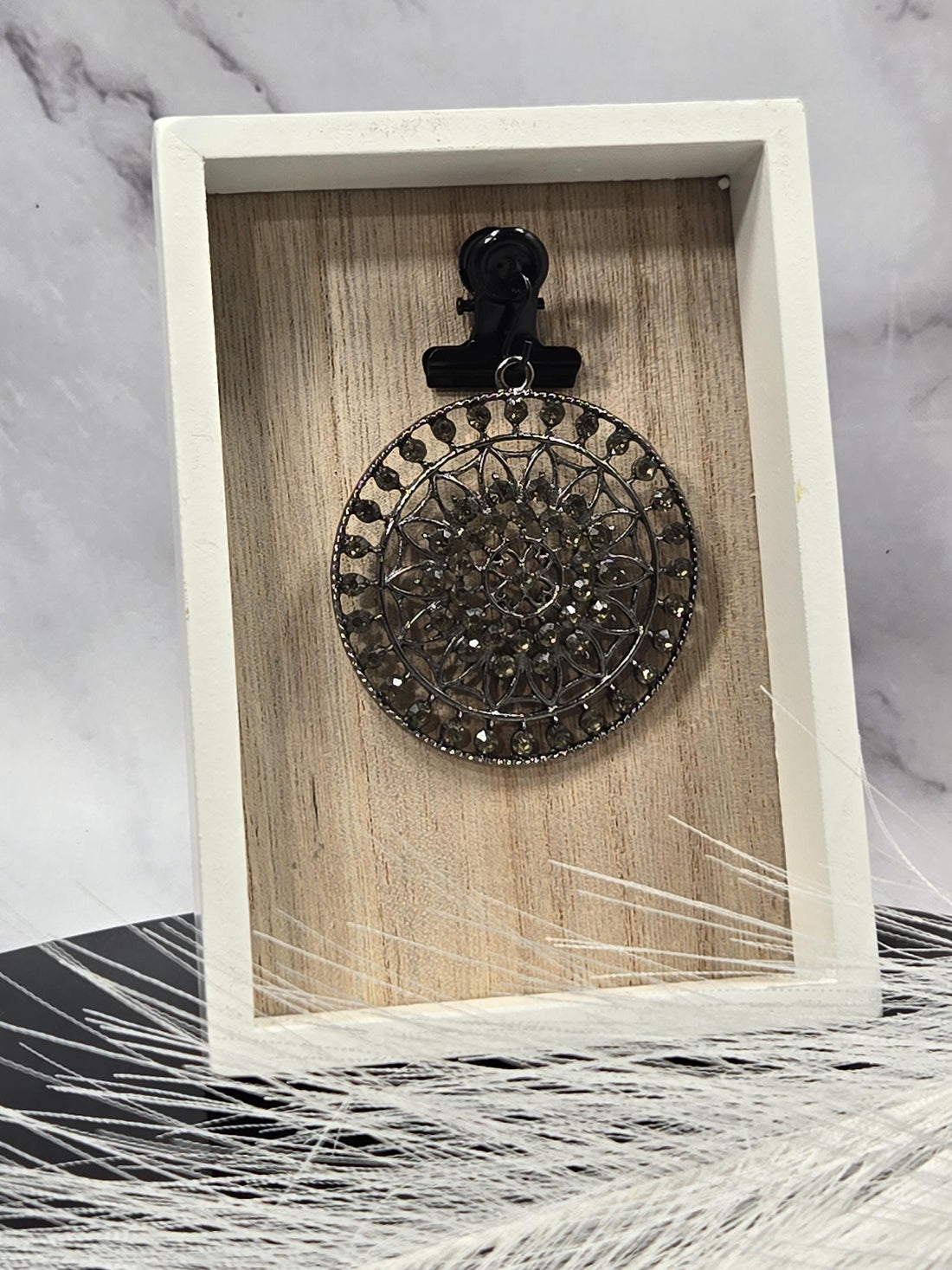 A decorative black metal piece with a circular pattern is displayed in a white wooden box against a marble background.