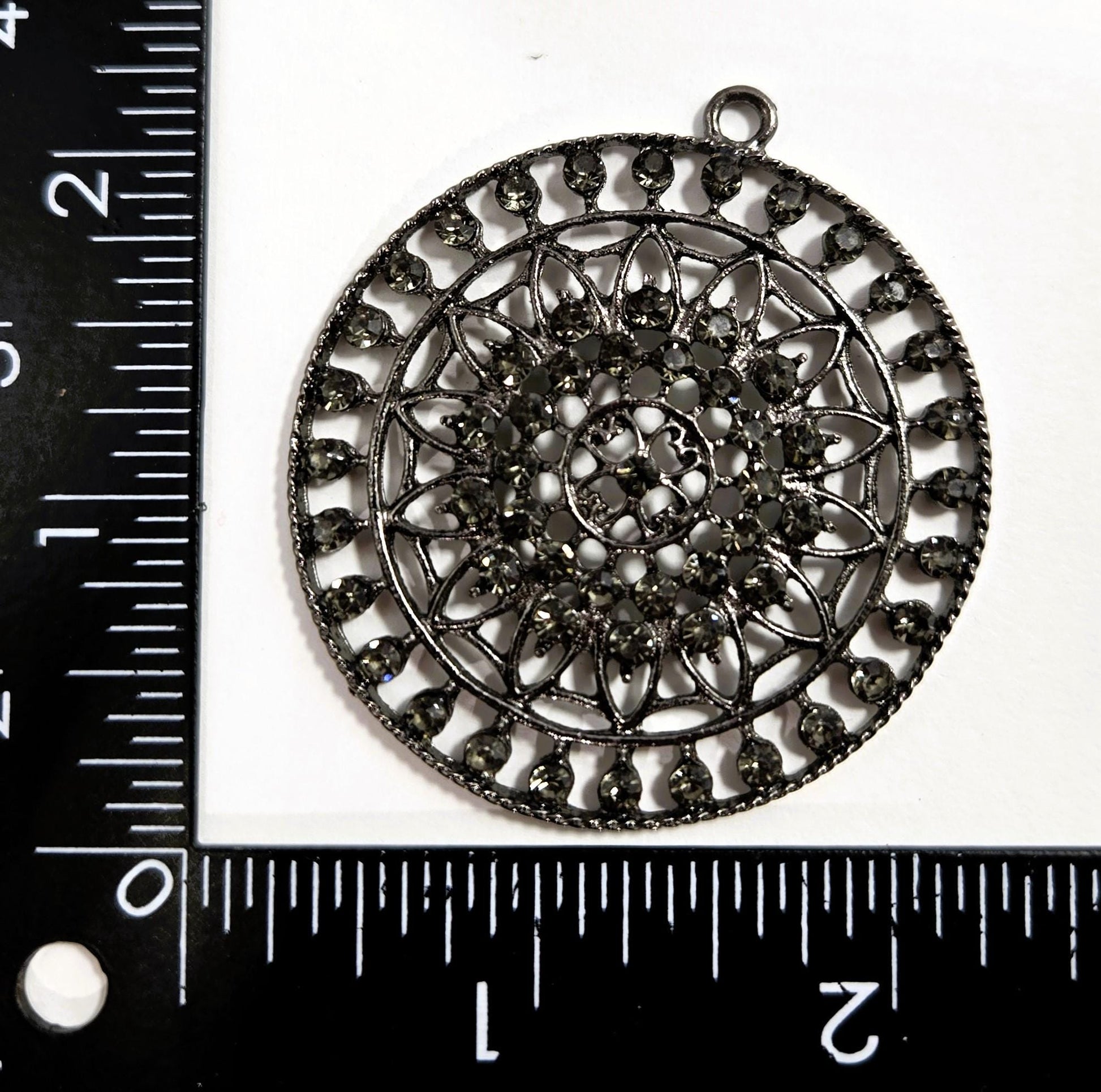 a large, intricately designed metal pendant with a floral pattern. The pendant is placed next to a ruler, which is used to measure its size.