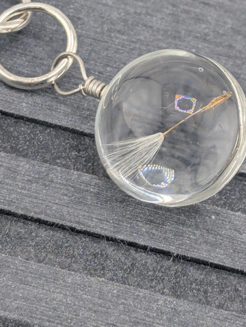 A clear glass keychain with a dandelion seed attached to it, resting on a wooden surface.