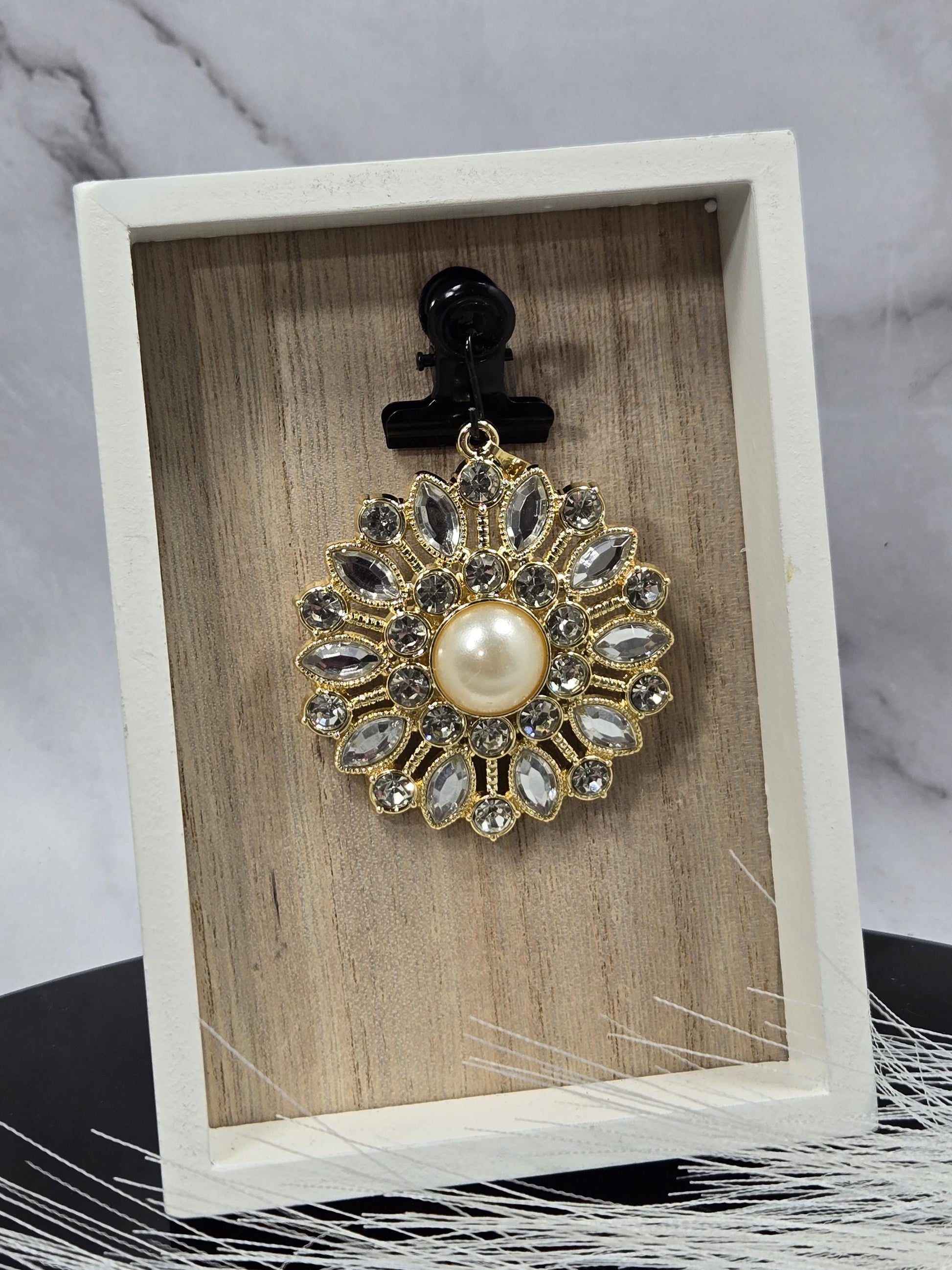 A decorative brooch with a pearl at its center, surrounded by numerous sparkling gemstones, is displayed in a white wooden box.