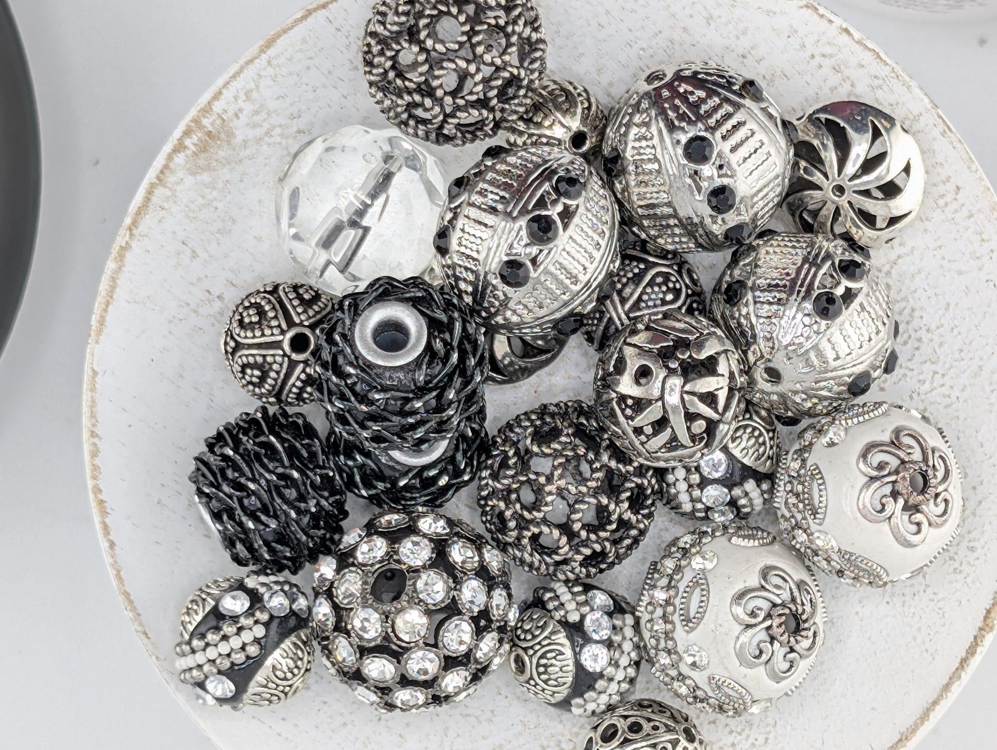 A white plate filled with various decorative beads and charms, including some with intricate designs and sparkling embellishments.