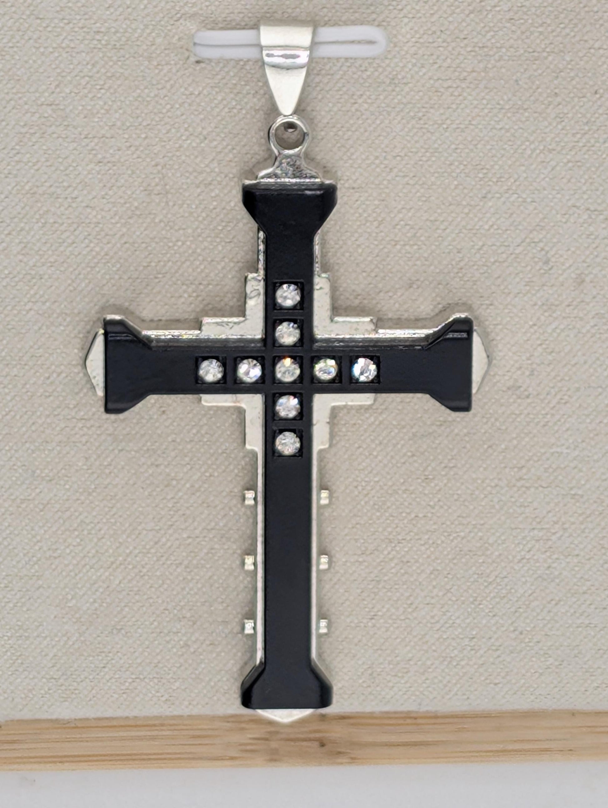 A silver-toned cross pendant with black accents and small rhinestones or crystals embedded in it, hanging against a beige background.