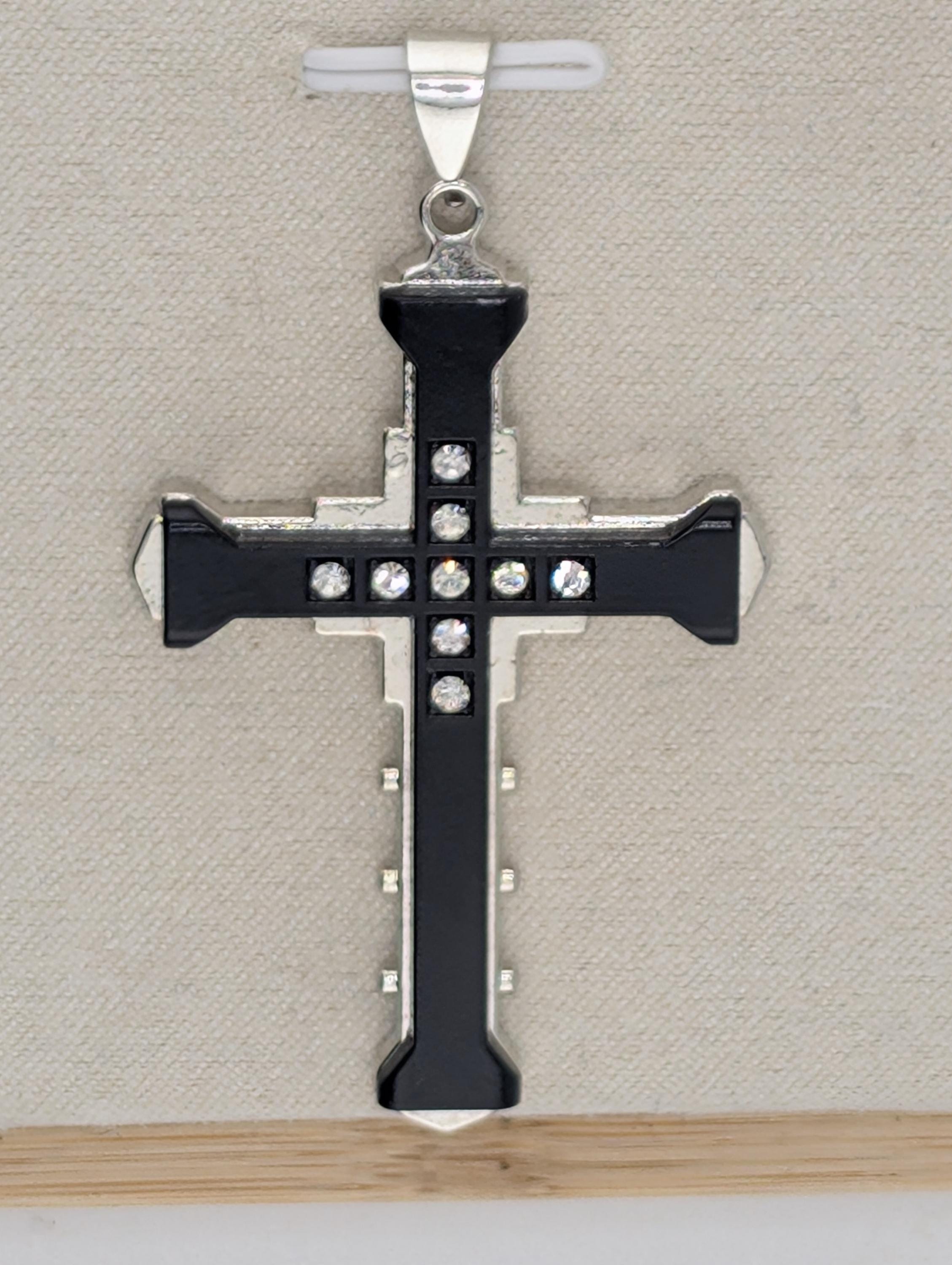 A silver-toned cross pendant with black accents and small rhinestones or crystals embedded in it, hanging against a beige background.
