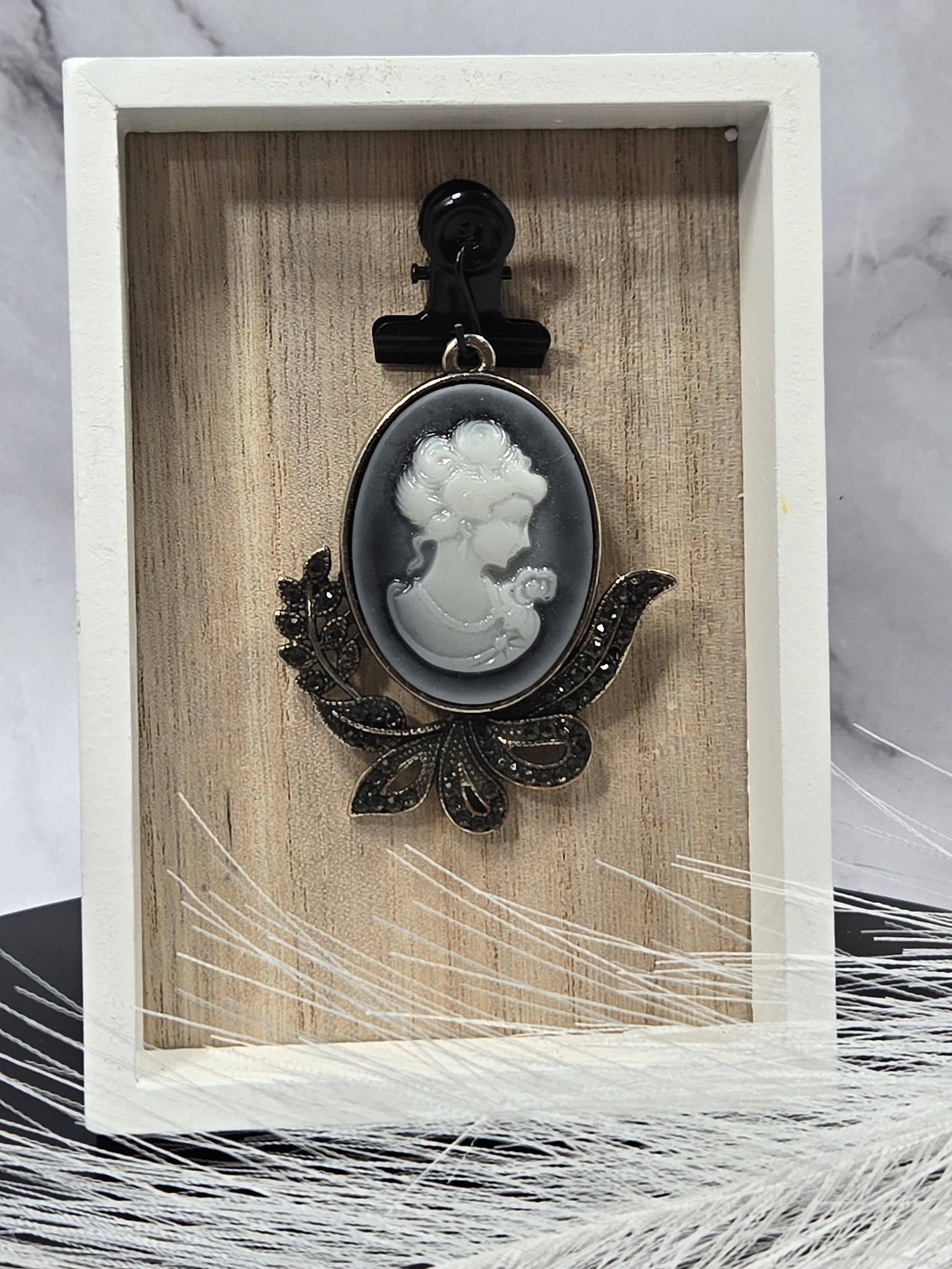 A white porcelain brooch with a black floral design is displayed in a white wooden box.