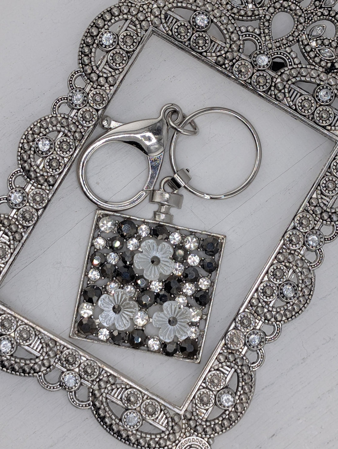 a silver-colored keychain with a decorative metal frame, adorned with small, clear rhinestones or crystals.