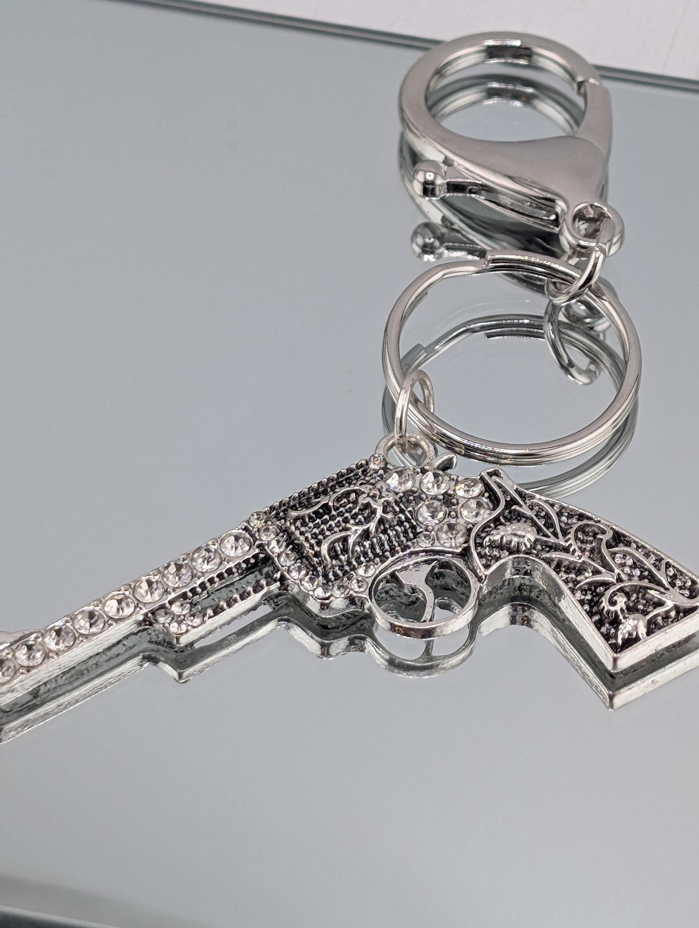 A silver-colored keychain with a detailed design, including a handle and a trigger mechanism, is displayed on a reflective surface.