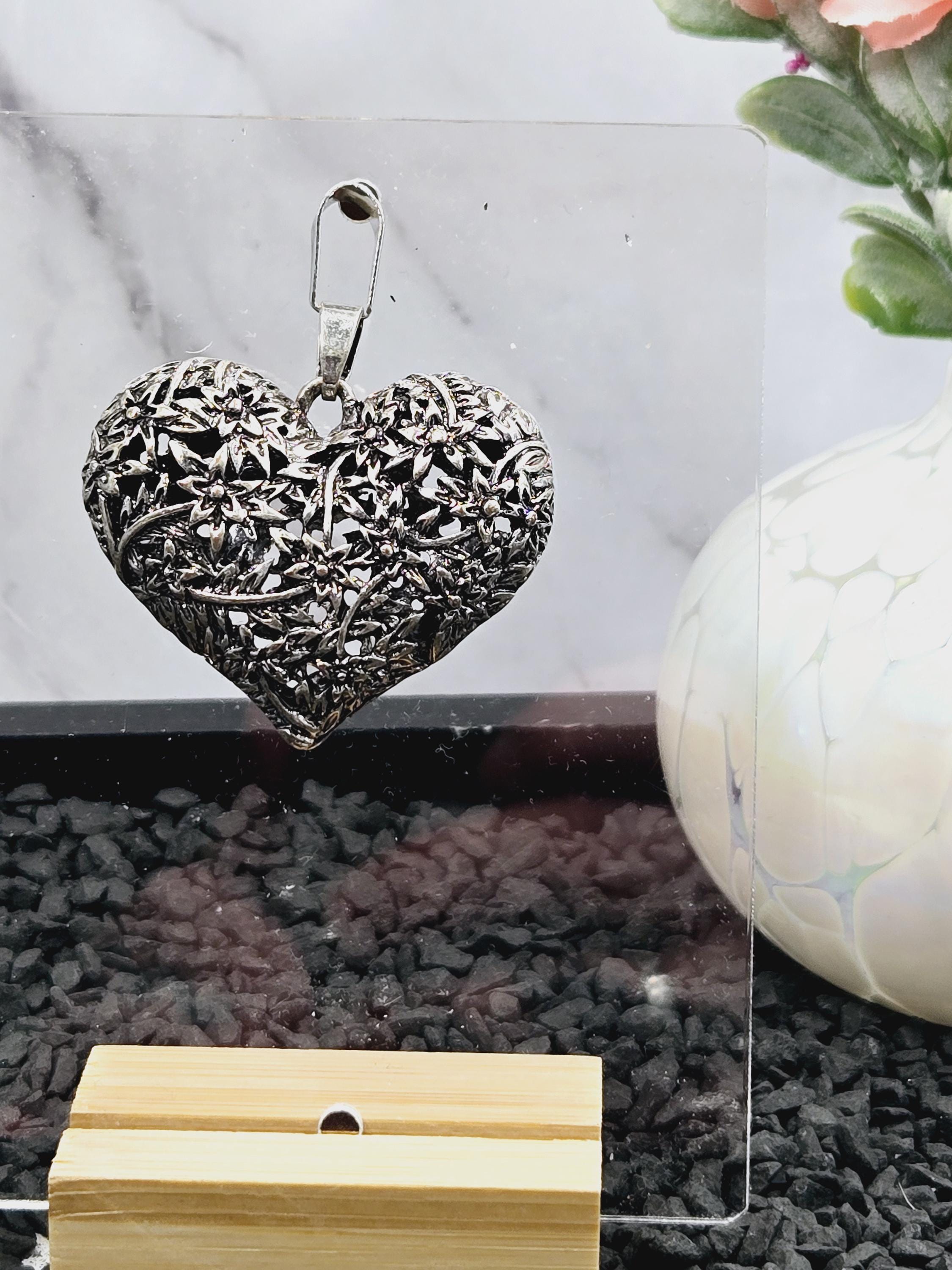a heart-shaped metal object with a floral design, placed on a marble surface next to a white vase and a wooden object.