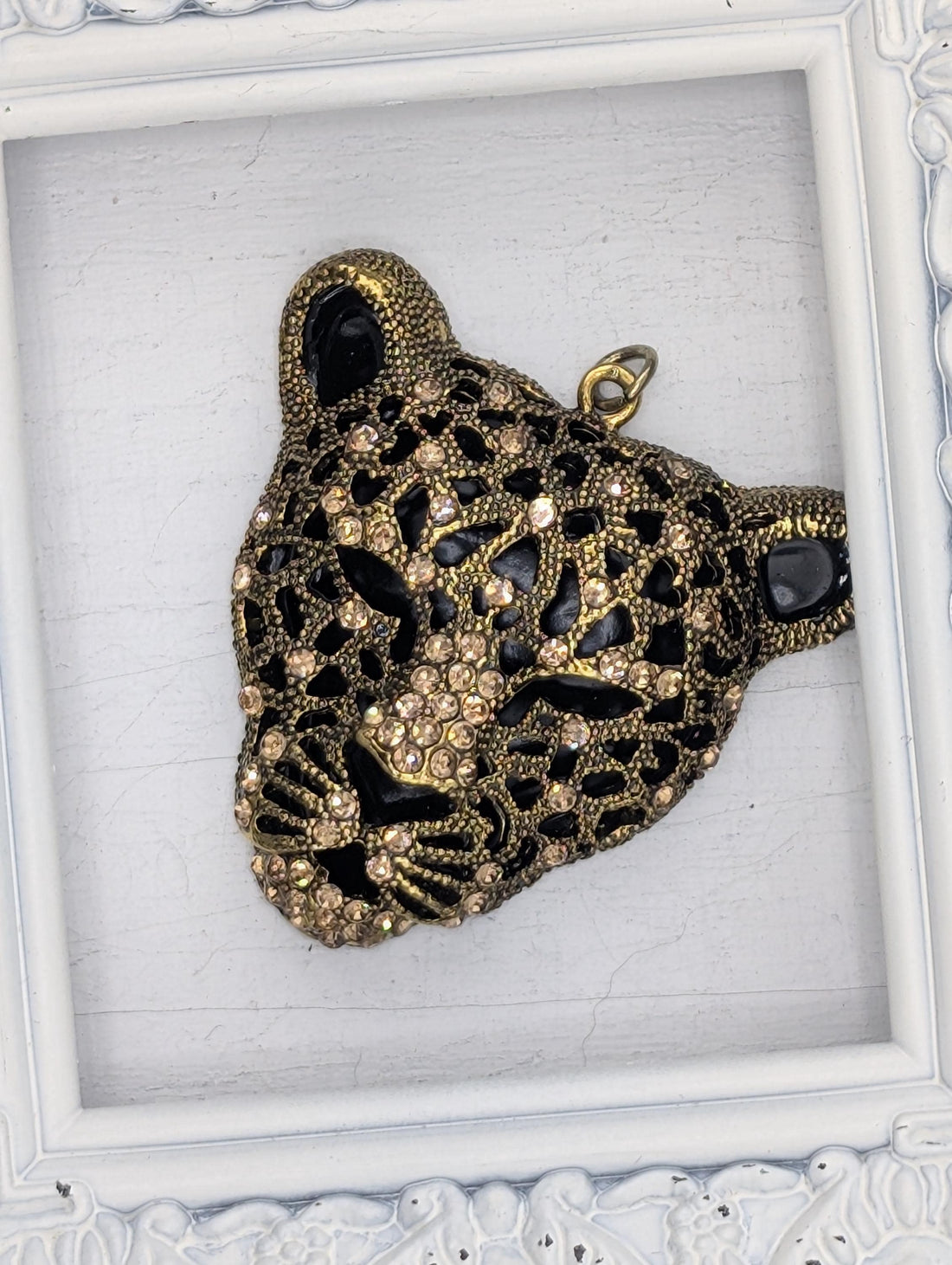 A decorative pendant in the shape of a leopard's head, adorned with intricate gold and black designs and sparkling rhinestones.