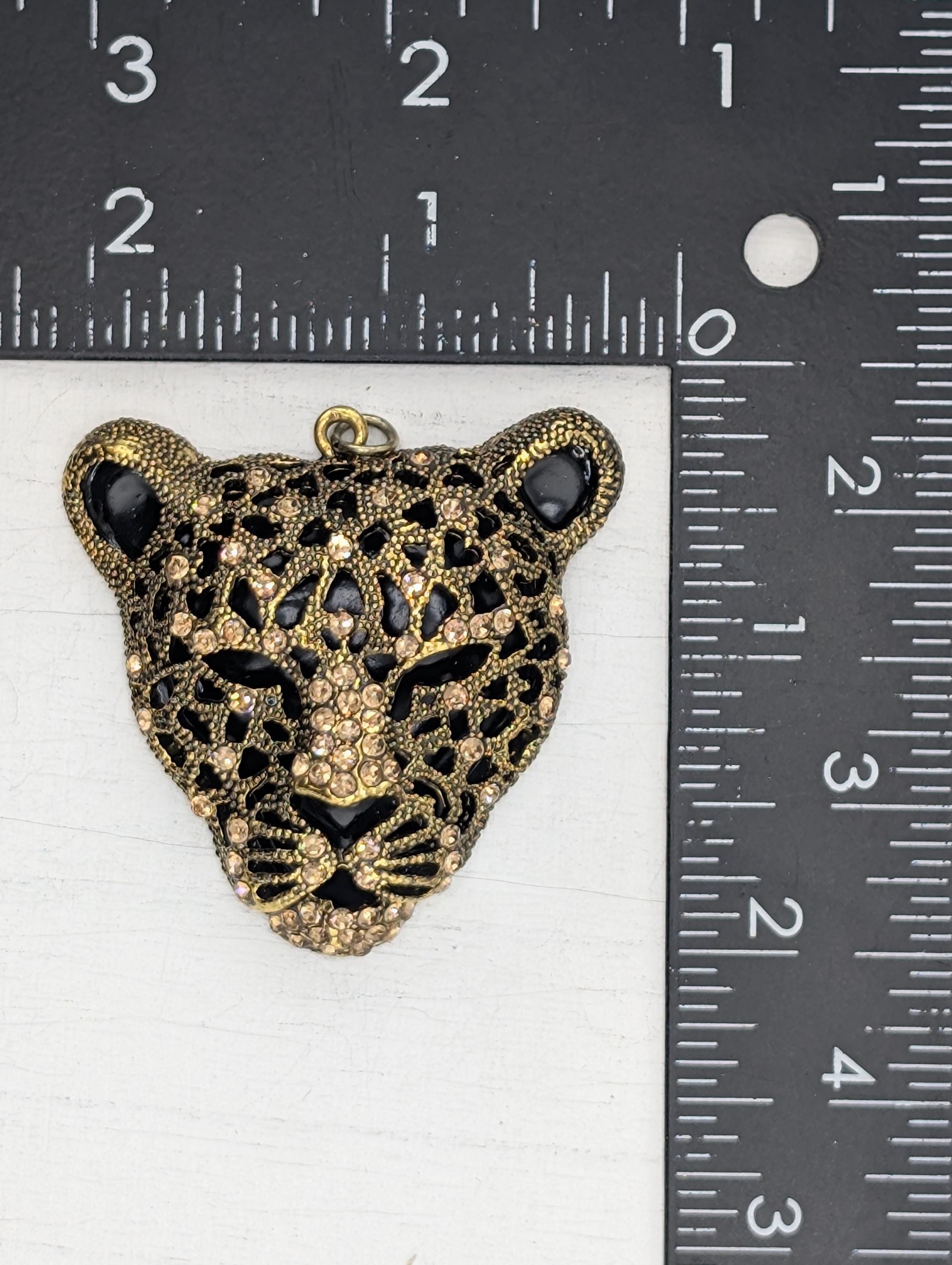 A close-up image of a decorative pendant in the shape of a leopard's head, adorned with rhinestones and gold accents.