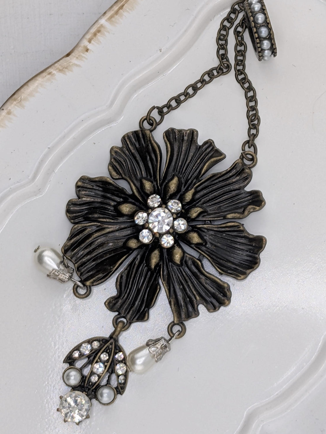 A decorative piece of jewelry featuring a large, intricately designed flower-shaped pendant with a central stone surrounded by smaller stones. The pendant is attached to a chain with pearls hanging from it, creating a stylish and elegant accessory.