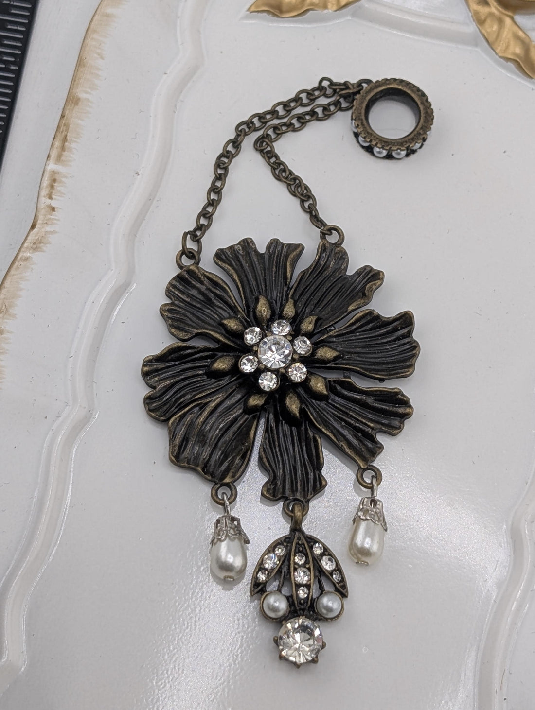 A decorative piece of jewelry featuring a large, intricately designed flower-shaped pendant with a central stone surrounded by smaller stones. The pendant is attached to a chain with pearls hanging from it, creating a stylish and elegant accessory.