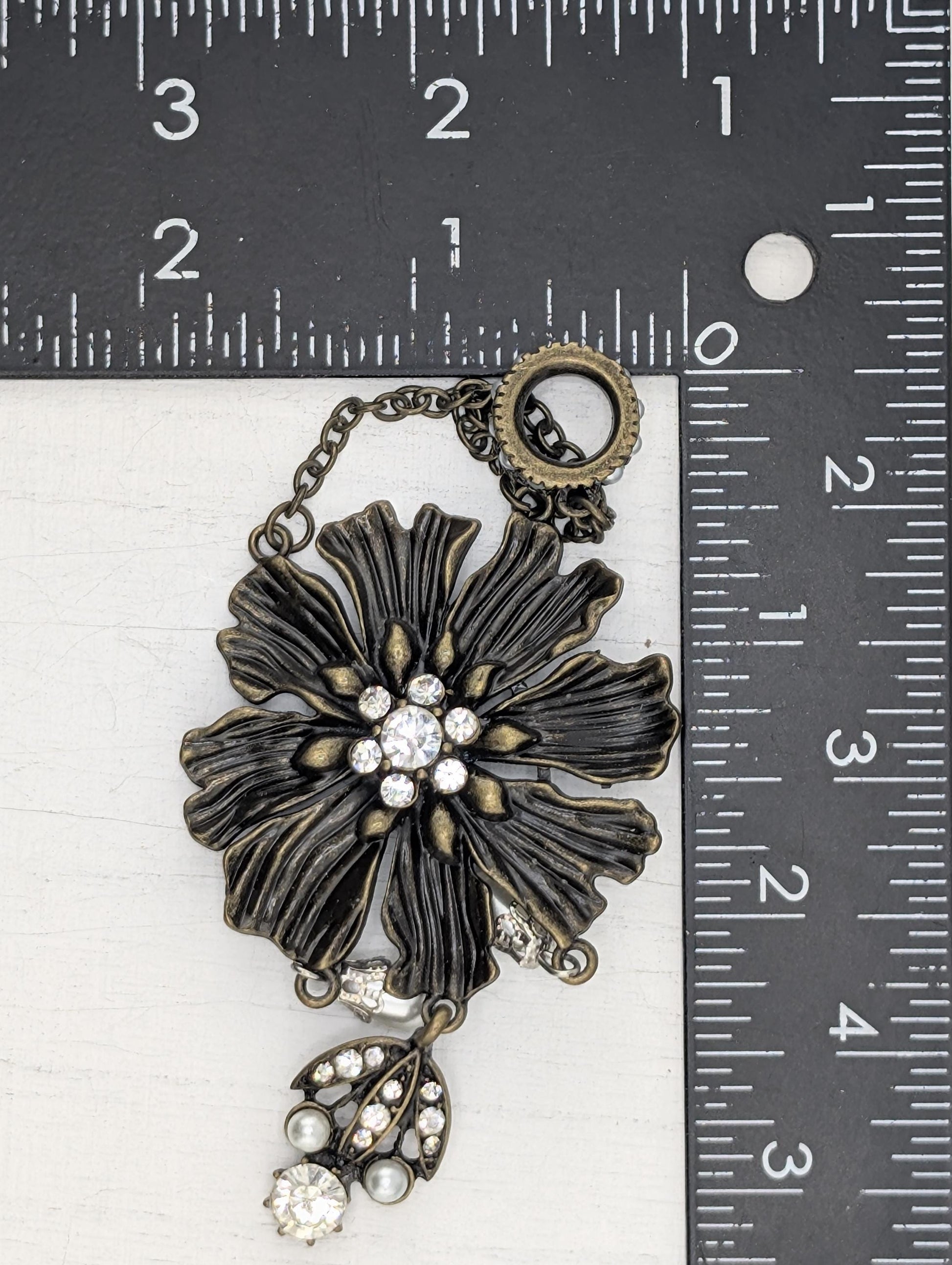 A close-up image of a decorative piece with a large, intricate flower-like design and sparkling rhinestones or crystals. The piece is placed next to a ruler for size comparison.