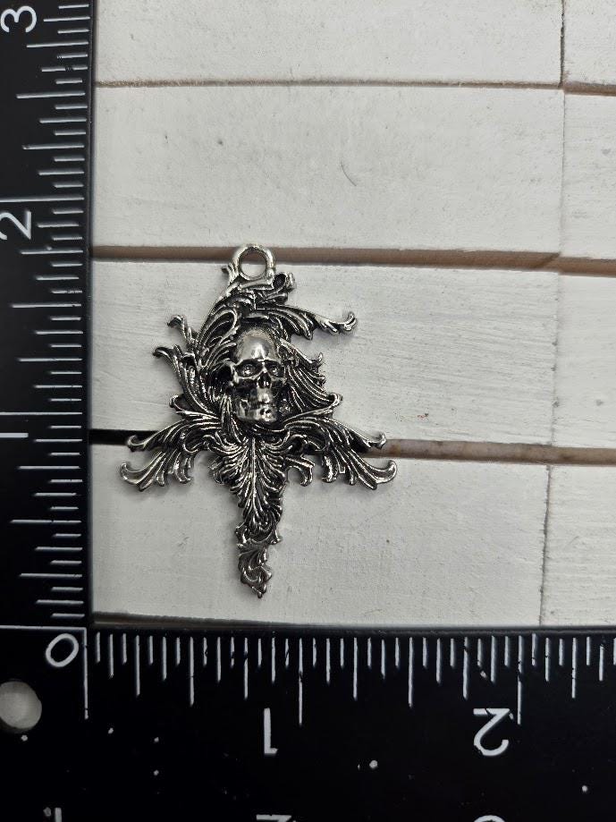 A silver-colored metal charm or pendant with an intricate design, featuring a skull or skull-like figure surrounded by ornate, decorative elements. The charm is placed next to a ruler for size reference.