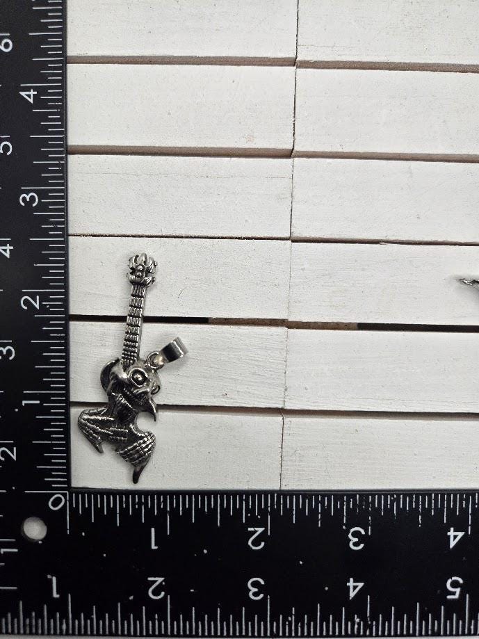 A silver-colored pendant in the shape of a guitar is placed next to a ruler on a white wooden surface.