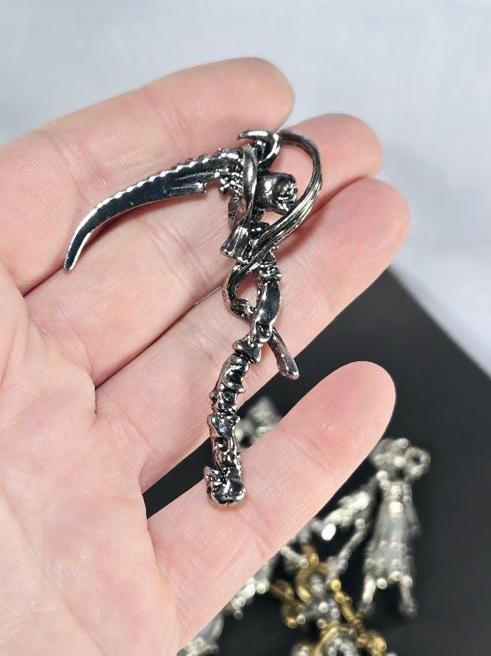 A hand holding a silver-colored metal pendant or charm in the shape of a skull or skeleton.