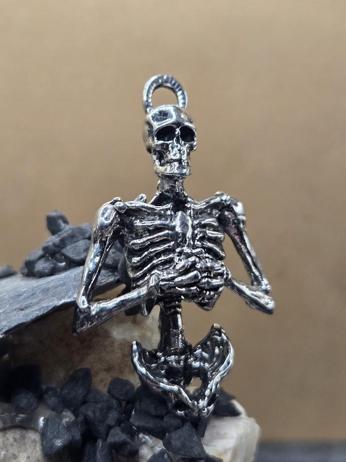 A silver-colored metal skeleton pendant is displayed against a beige background.