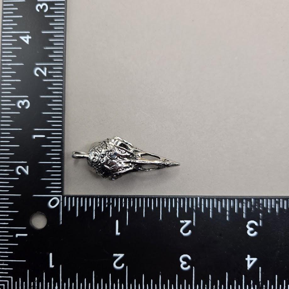 A silver-colored metal object, possibly a pendant or brooch, is placed next to a ruler on a white surface.