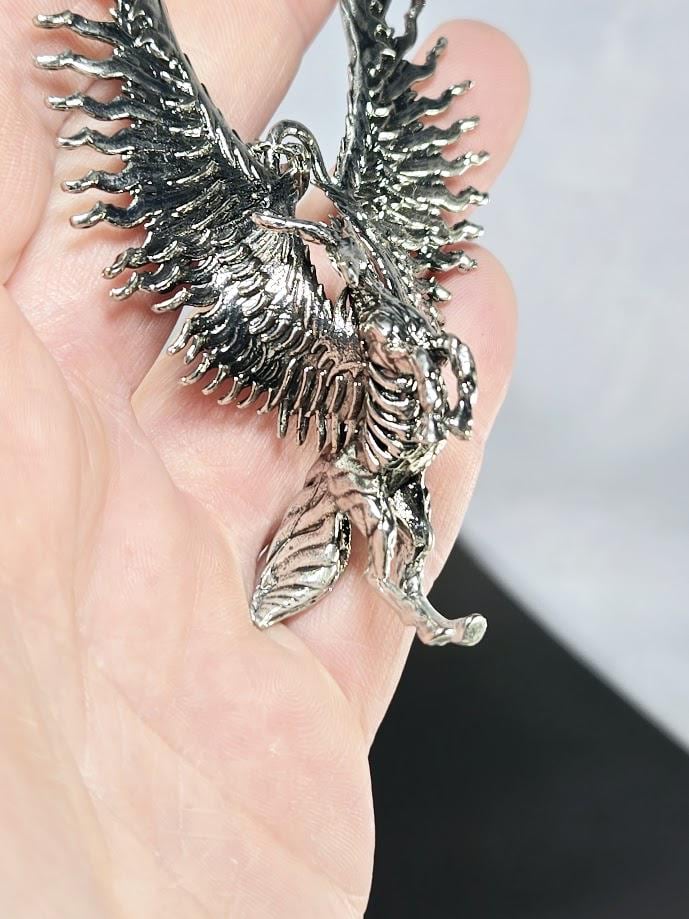 A close-up image of a person's hand holding a silver-colored metal pendant in the shape of a bird or winged creature.