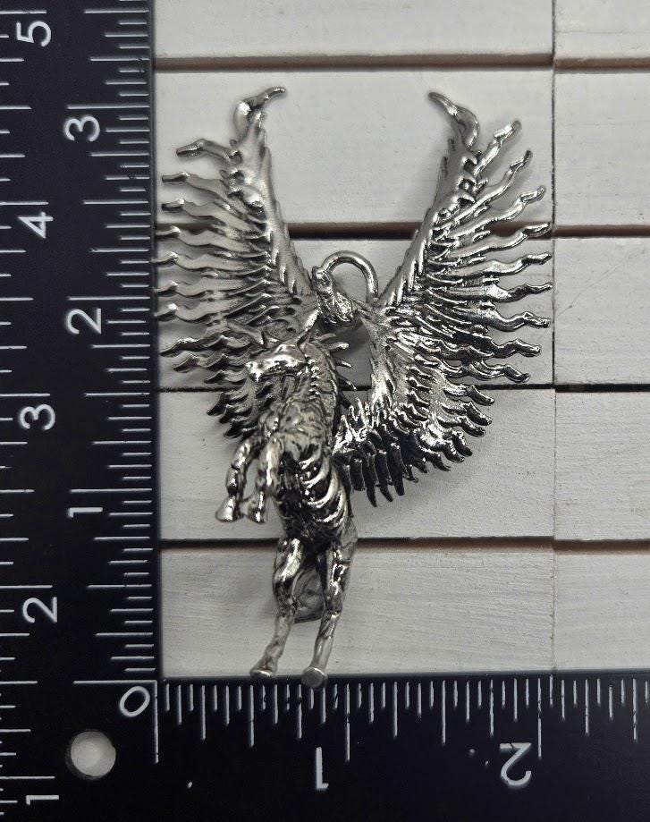 A silver-colored metal pendant in the shape of a winged creature, possibly a mythical or fantastical animal, is being measured against a ruler.