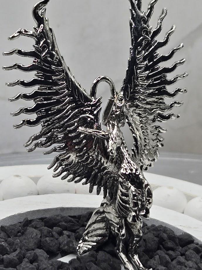 A metallic, winged sculpture with intricate details and a metallic sheen, displayed on a bed of small rocks.