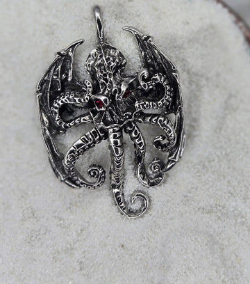 A silver pendant in the shape of a winged creature with intricate details and a red gemstone.