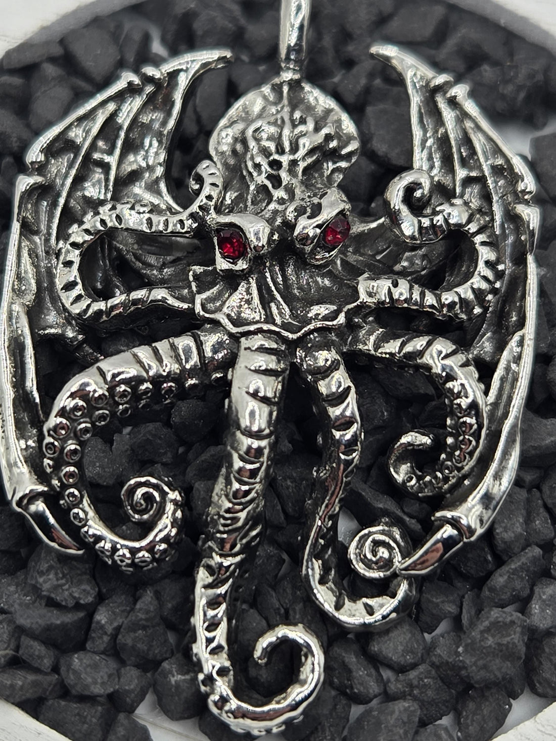 A silver pendant in the shape of a winged creature with intricate details and a red gemstone.