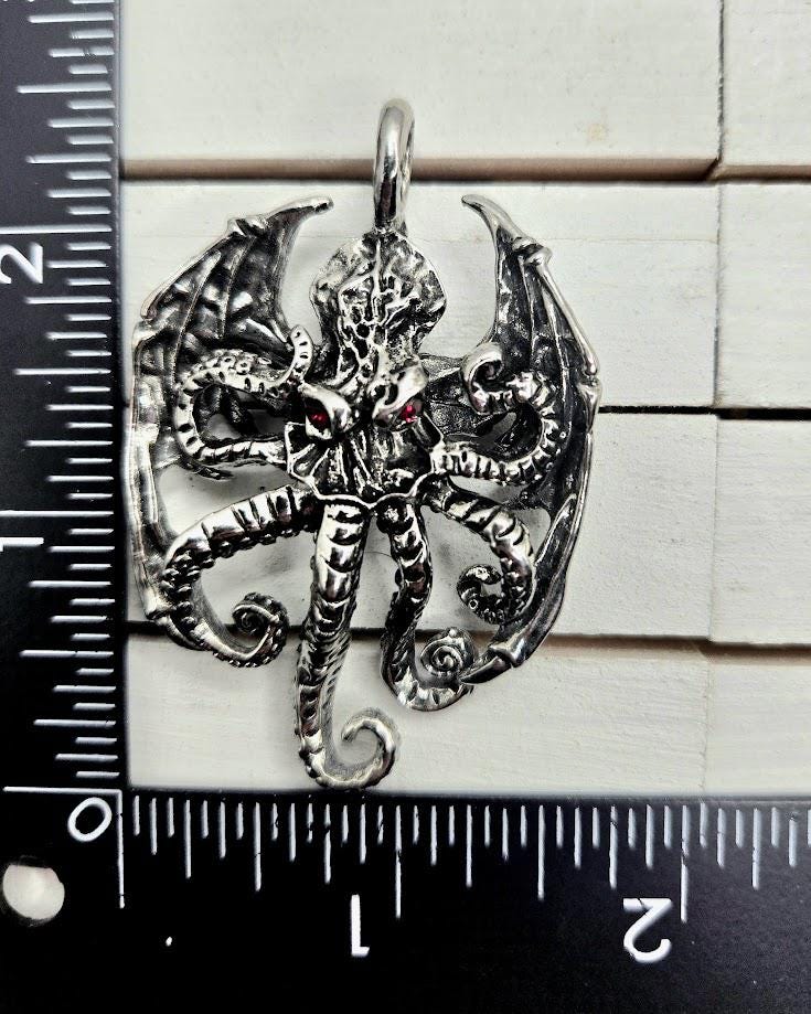 A silver pendant in the shape of a tentacles creature with red eyes, displayed against a ruler for size reference.