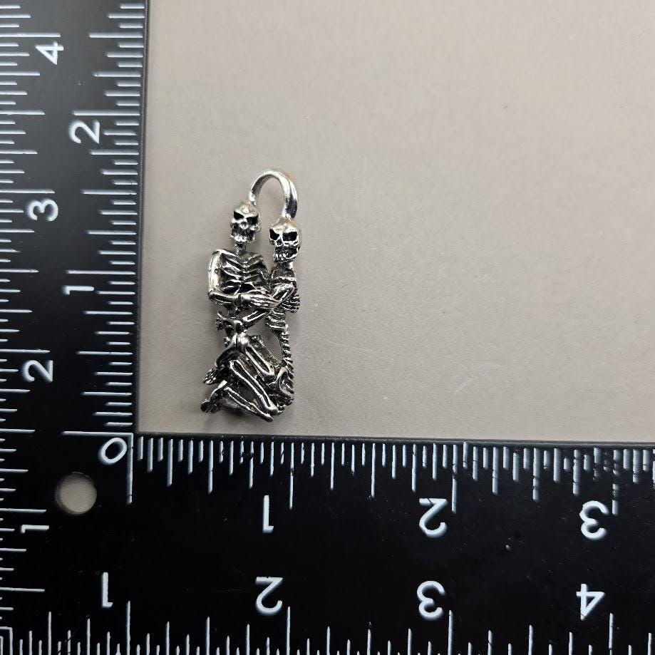 A silver-colored metal charm or pendant in the shape of a skeleton is placed next to a ruler on a black surface.