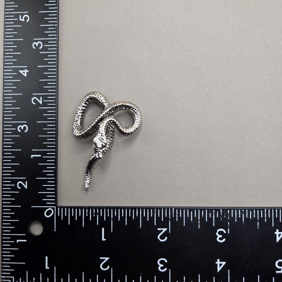 A silver snake brooch is placed next to a ruler on a gray surface.
