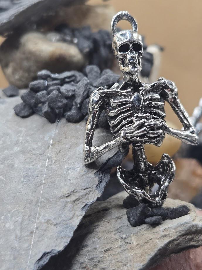 A silver-colored metal skeleton pendant is displayed on a rock, surrounded by small rocks and pebbles.