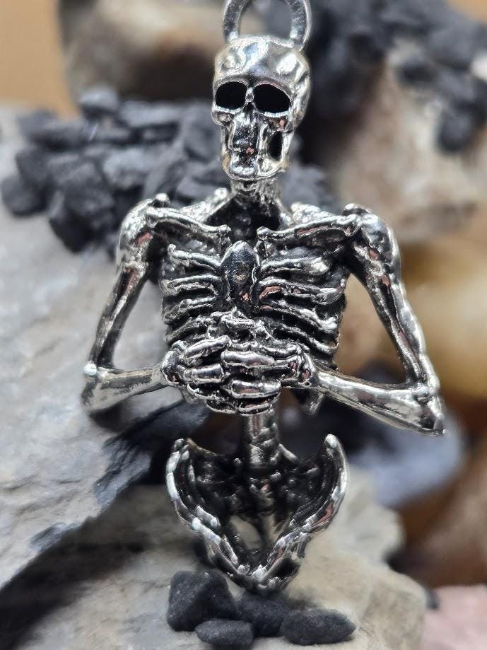 A silver-colored metal skeleton pendant is displayed against a blurred background of rocks and stones.