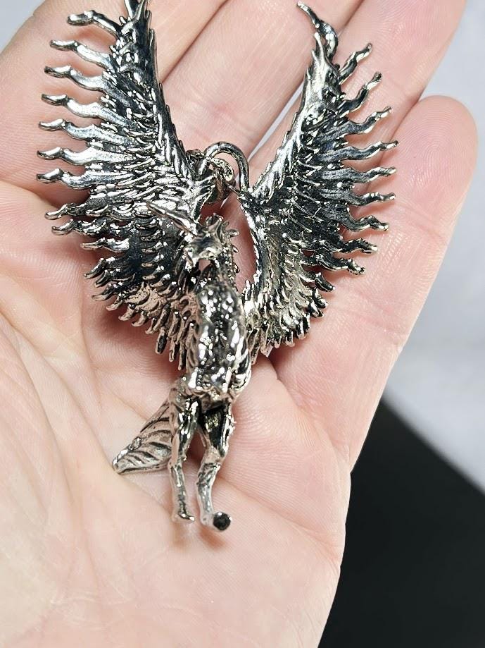A hand holding a silver-colored pendant in the shape of a winged creature, possibly a mythical or fantastical animal.