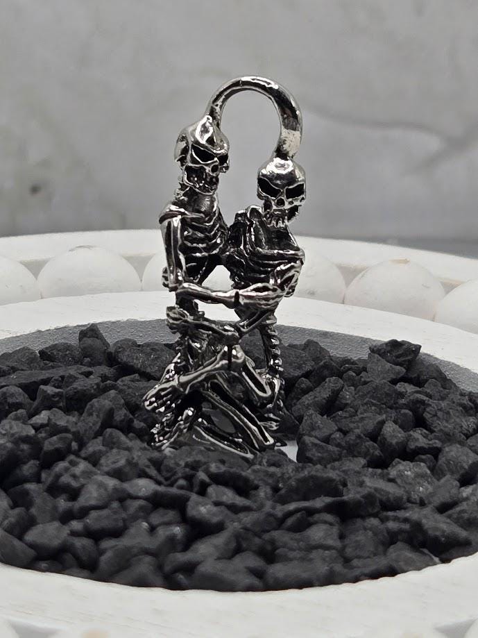A silver metal sculpture of two skeletal figures embracing each other, set against a background of small black rocks.