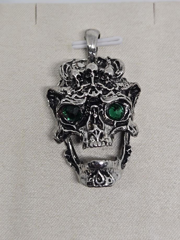 A silver-colored pendant in the shape of a skull with green gemstones on the eyes.