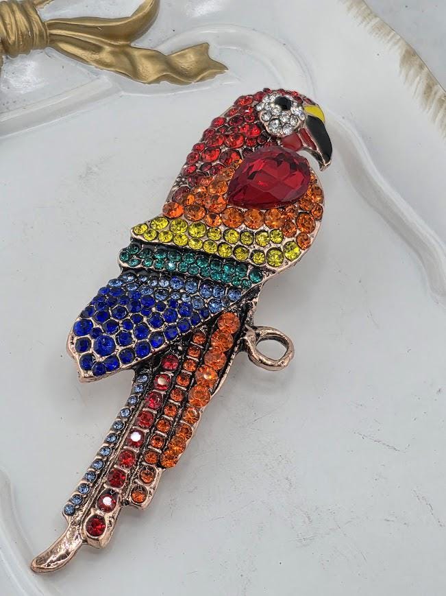 A colorful, ornate bird brooch made of various materials, including gemstones and metal, is displayed on a white surface.