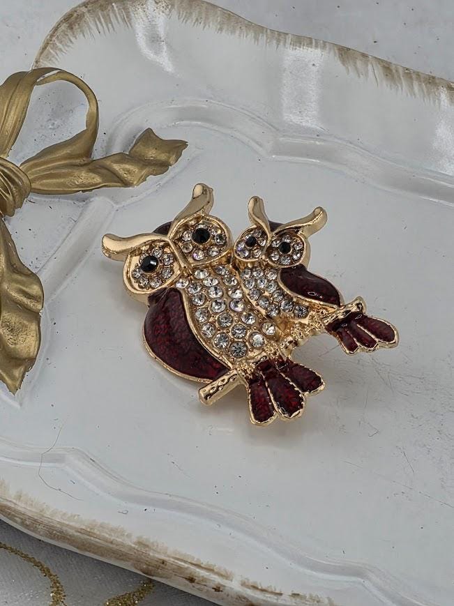 A decorative brooch in the shape of two owls, with one owl being larger and the other smaller, is displayed on a white tray.