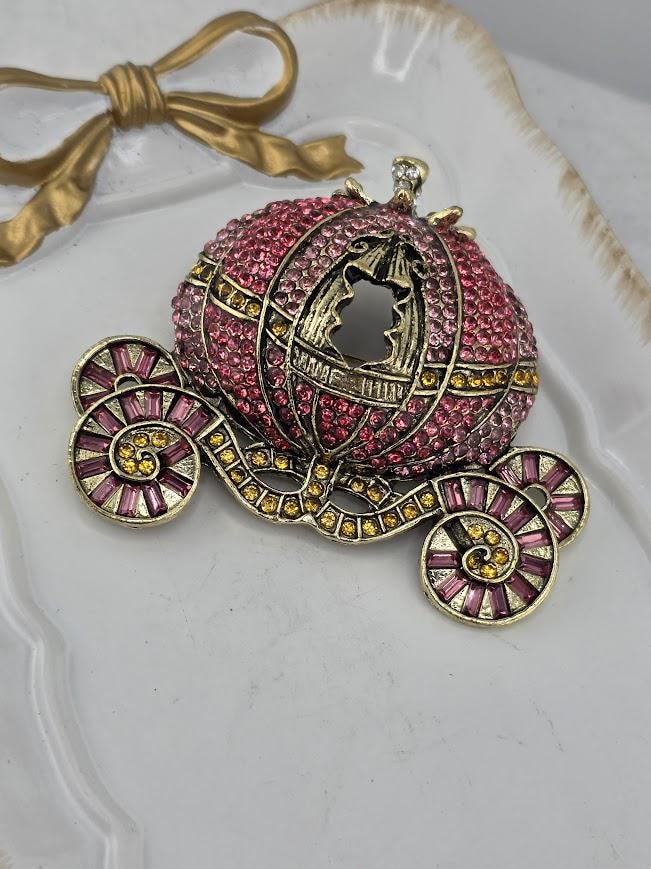 A decorative piece featuring a large, ornate carriage-shaped brooch with a pink and gold color scheme, adorned with rhinestones and a gold bow.