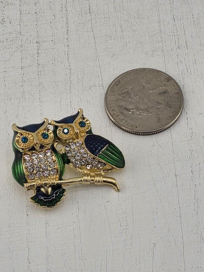 A decorative brooch in the shape of two owls, with one owl wearing a green and blue color scheme and the other in gold and blue, is placed next to a United States quarter.