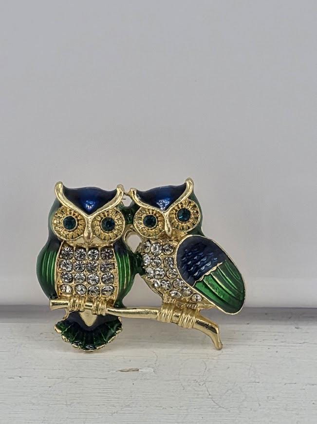 A decorative brooch featuring two ornate owls, one with green and blue accents and the other with blue and gold accents, sitting on a branch.