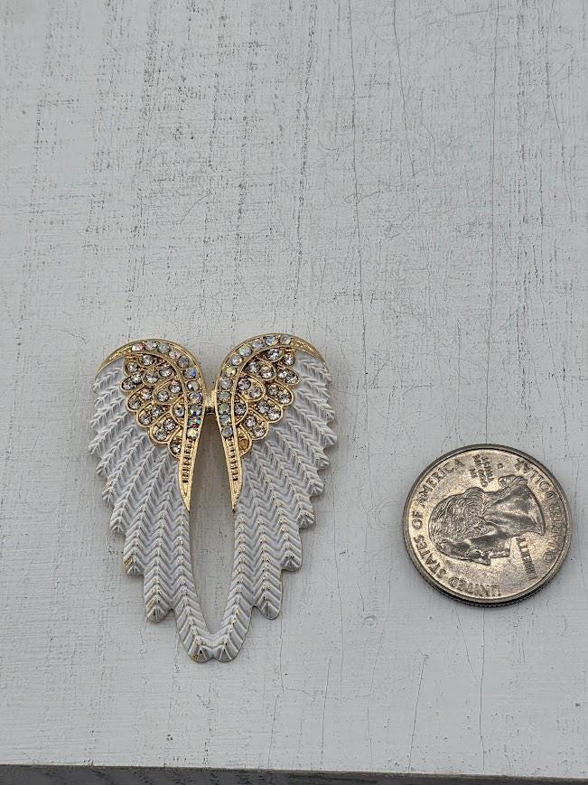 A decorative white and gold wing brooch with rhinestones is displayed against a black background, framed by a white ornate frame.