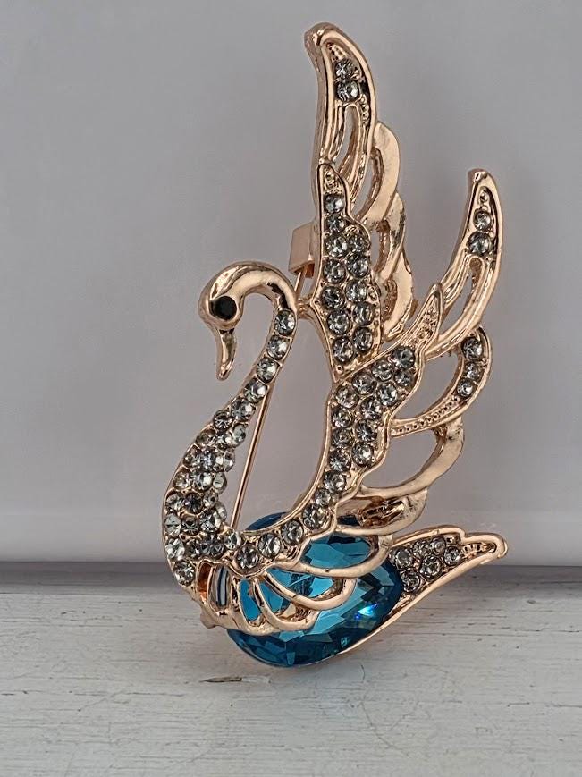 A decorative gold-colored pin or brooch in the shape of a swan, adorned with sparkling rhinestones or crystals.