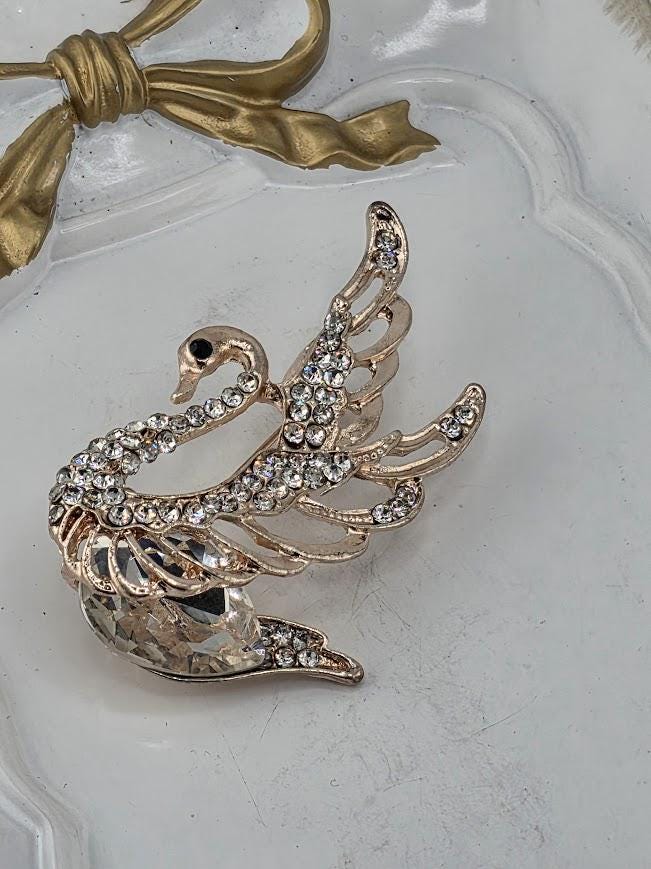A decorative gold-colored swan brooch with rhinestones or crystals on its wings and body, placed on a white surface.