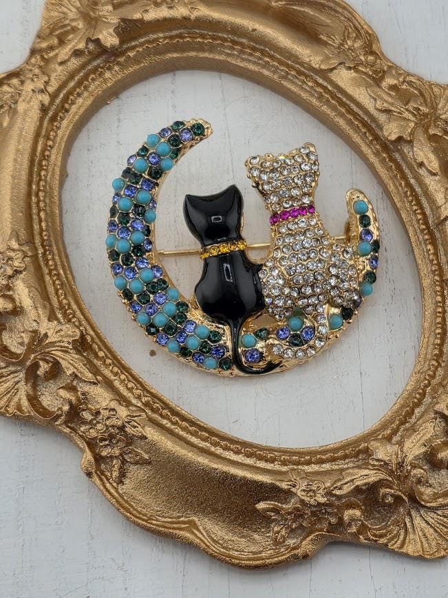A decorative brooch in the shape of a crescent moon, featuring two cats - one black and one white - sitting on the moon. The brooch is surrounded by a gold frame with intricate designs.