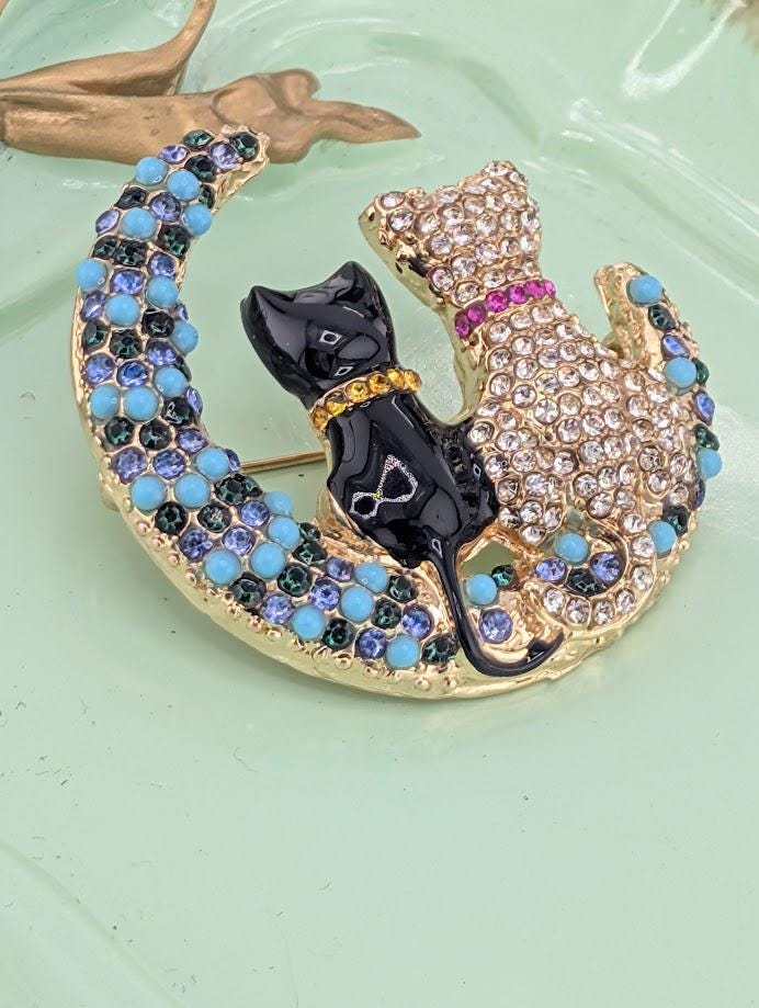 A decorative brooch featuring a black cat and a gold cat on a crescent moon, with blue and purple gemstones.