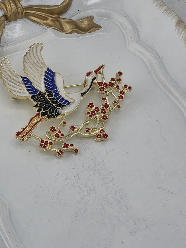 A decorative brooch with a crane and flowers, featuring a combination of blue, red, and gold colors.