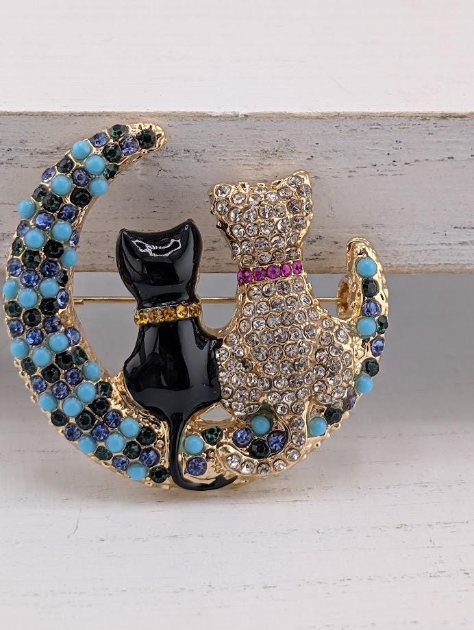 A decorative brooch featuring two cats, one black and one white, sitting on a crescent moon. The cats are adorned with gemstones and the moon is embellished with blue and green stones.