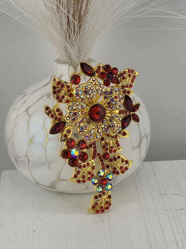 A decorative brooch with a floral design and red gemstones is displayed on a white vase.