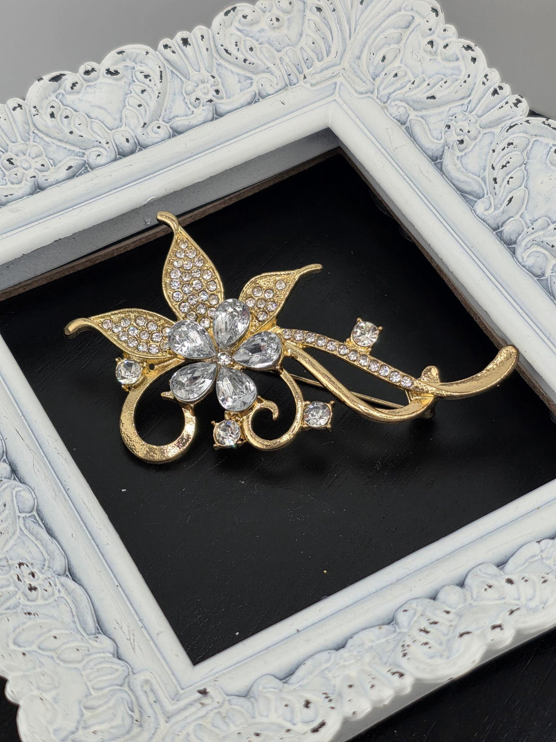 A decorative brooch with a floral design and sparkling gemstones is displayed in a white ornate frame.