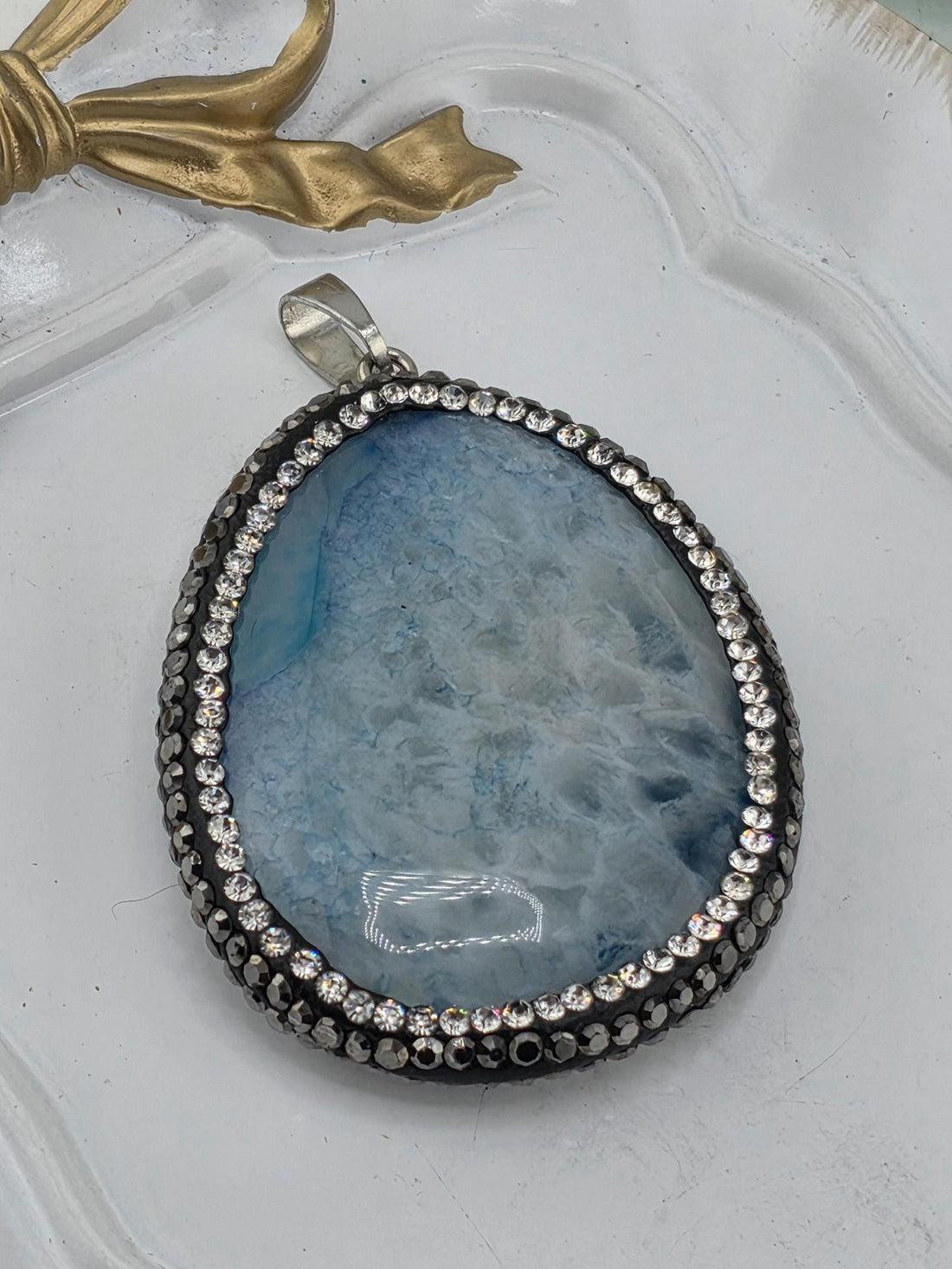 A large, blue-green stone pendant with a silver frame and sparkling rhinestones, resting on a white surface.